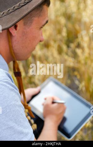 Digitale Farm. Porträt eines Bauern, der im goldenen Weizenfeld sitzt und auf dem Tablet scrollt. Junger Mann mit Cowboyhut auf dem Feld, der Weizenernte untersucht. Hafer Stockfoto