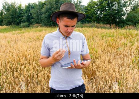 Junger Bauer. Porträt eines Bauern, der im goldenen Weizenfeld sitzt und auf dem Tablet scrollt. Junger Mann mit Cowboyhut auf dem Feld, der Weizenernte untersucht. Hafer Stockfoto