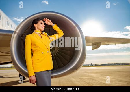 Fröhliche Angestellte der Fluggesellschaft, die mit der Hand die Augen vor der Sonne verdeckte und lächelte, während sie in der Nähe des Triebwerks der Flugzeugturbine stand Stockfoto