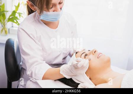 Kosmetologe in medizinische Maske und Schutzhandschuhe machen Verjüngung Verfahren in der Kosmetologie Klinik zu einer schönen lächelnden Frau. Mechanisch Stockfoto