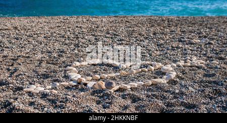 Liebe Herzform auf Sand aus weißen Kieselsteinen, Symbol der Liebe am Strand mit Meereshintergrund. Valentinstag Liebe Symbol der Herzform, Liebe Stockfoto