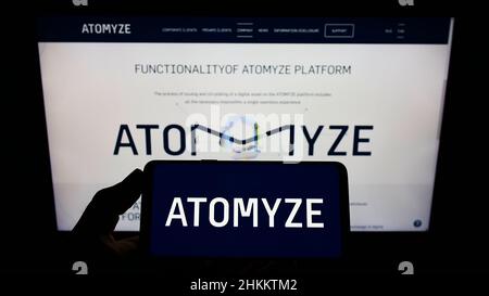 Person, die Mobiltelefon mit Logo des Schweizer Technologieunternehmens ATOMYZE AG auf dem Bildschirm vor der Business-Webseite hält. Konzentrieren Sie sich auf die Telefonanzeige. Stockfoto