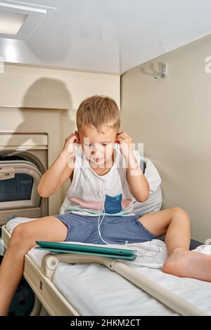 Blonde kurzhaarige kleine Junge in legerer Kleidung sieht Cartoons über Tablet-PC mit Kopfhörern sitzen auf dem oberen Regal in der Bahn Auto Stockfoto