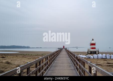 Kleiner rot-weißer Leuchtturm aus Holz an einem winterlichen Sandstrand neben einem hölzernen Steg ohne Menschen. Stockfoto