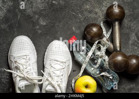 Sporthintergrund Draufsicht auf Sneakers, Hanteln, Wasserflasche, Apfel und Höhenmesser-Band auf schwarzem Hintergrund mit Kopierfläche. Gesunder Lebensstil Stockfoto