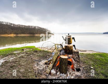 Türkische traditionelle Teekannen kochten am Lagerfeuer, ein glatter See im Hintergrund. Stockfoto