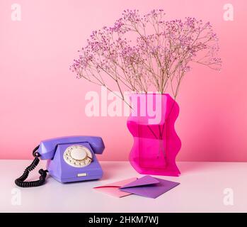 Retro-Telefon am Tisch mit getrockneten Blumen in Vase und Umschlägen. Set von Objekten auf rosa Hintergrund. Valentinstag-Konzept. Hochwertige Fotos Stockfoto
