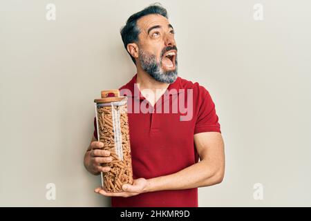 Mann mittleren Alters mit Bart und grauen Haaren hält Glas mit Makkaroni Pasta wütend und verrückt Schreien frustriert und wütend, Schreien mit Wut aufschauen Stockfoto