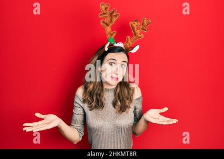 Junge hispanische Mädchen trägt Hirsch weihnachtshut ahnungslos und verwirrt Ausdruck mit Armen und Händen angehoben. Zweifel Konzept. Stockfoto