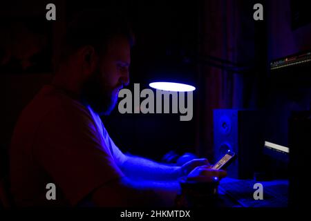 Ein Mann mit Bart und Telefon sitzt nachts an einem Tisch gegenüber einem Monitor in einem Raum. Stockfoto