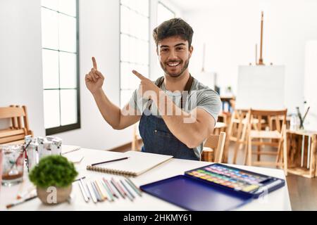 Junger hispanischer Mann im Kunststudio lächelt und blickt auf die Kamera, die mit zwei Händen und Fingern zur Seite zeigt. Stockfoto