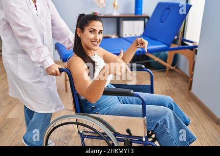Junge hispanische Frau, die auf dem Rollstuhl in der Physiotherapie-Klinik sitzt, lächelt glücklich und zeigt mit Hand und Finger Stockfoto