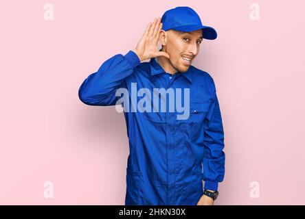 Glatze Mann mit Bart tragen Baumeister Jumpsuit Uniform lächeln mit Hand über Ohr hören ein Hören Gerücht oder Klatsch. Taubheit Konzept. Stockfoto