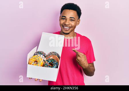 Junger afroamerikanischer Mann hält Box mit leckeren bunten Donuts lächelnd glücklich zeigt mit Hand und Finger Stockfoto