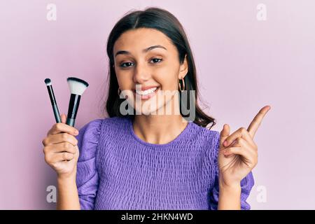 Junge hispanische Frau hält Make-up Bürsten lächelnd glücklich zeigt mit Hand und Finger zur Seite Stockfoto