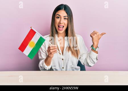 Junge hispanische Frau mit der Flagge kurdistans auf dem Tisch sitzend, mit dem Daumen nach oben zur Seite zeigend, lächelnd mit offenem Mund Stockfoto