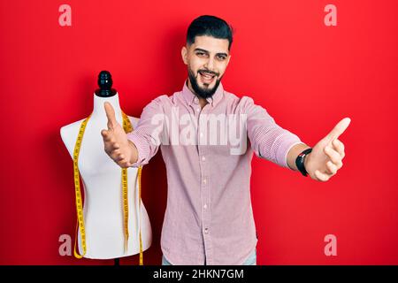 Hübscher Schneidermann mit Bart, der neben der Übungspuppe steht und die Kamera lächelt und die offenen Arme umarmt. Fröhlicher Ausdruck, der das Glück umarmt. Stockfoto