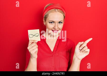 Junge blonde Frau hält unendliche Symbol Erinnerung lächelnd glücklich zeigt mit Hand und Finger zur Seite Stockfoto