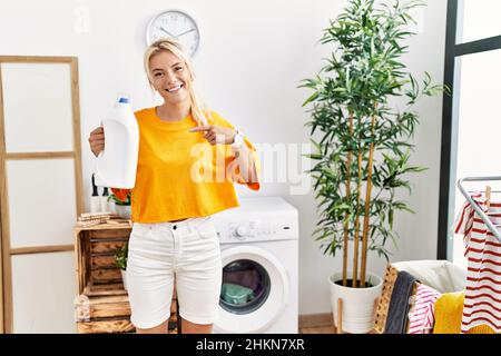 Junge kaukasische Frau, die ihre Wäsche waschen und die Waschmittelflasche lächelnd hält, zeigt glücklich mit Hand und Finger Stockfoto