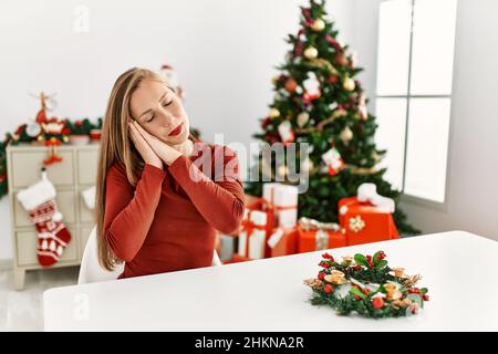 Kaukasische junge blonde Frau, die am weihnachtsbaum auf dem Tisch sitzt und müde schläft und mit den Händen zusammen posiert, während sie mit geschlossenem e lächelt Stockfoto