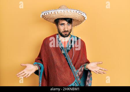 Junger hispanischer Mann mit mexikanischem Hut, ahnungsloser und verwirrter Expression mit erhobenen Armen und Händen. Zweifelskonzept. Stockfoto