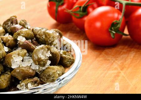 Gesalzene Kapern in einer Schüssel auf einem Schneidebrett. Rote Tomaten im Hintergrund. Stockfoto