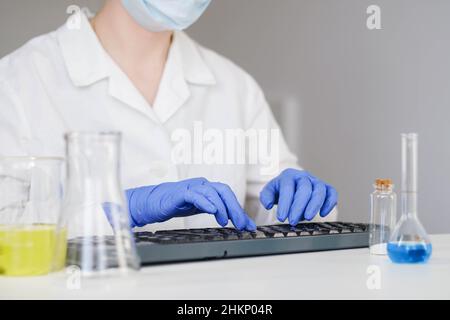 Wissenschaftlerin in Handschuhen, die im Labor auf der Tastatur am Schreibtisch arbeitet. Stockfoto