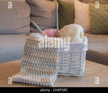 Rosa und weiße Wollkugeln in einem weißen Korb mit Stricknadeln und einem handgefertigten Strickschal Stockfoto