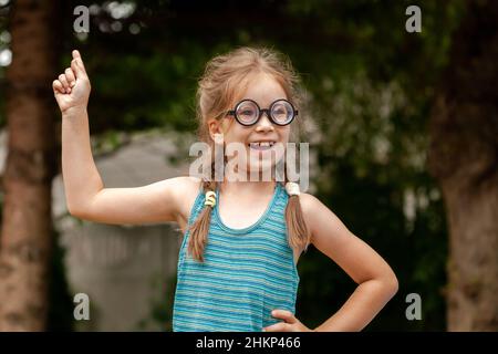 Junge glücklich neugierig Grundschulalter Mädchen, clever intelligent Genie Kind Schüler in großen lustigen Brille hat eine brillante Idee, Finger zeigt nach oben, Stockfoto