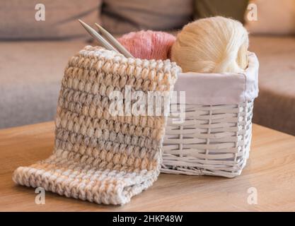 Rosa und weiße Wollkugeln in einem weißen Korb mit Stricknadeln und einem handgefertigten Strickschal Stockfoto