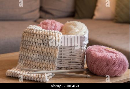 Rosa und weiße Wollkugeln in einem weißen Korb mit Stricknadeln und einem handgefertigten Strickschal Stockfoto