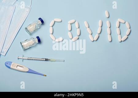 Die Inschrift kovid aus Pillen auf einem blauen medizinischen Hintergrund, neben einem Impfstoff, einer Spritze, einem Thermometer, einer Schutzmaske. Medizinisches Thema. Stockfoto