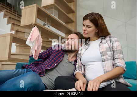 Das junge kaukasische Liebespaar ruht sich in Erwartung eines Babys auf einem Sofa im Wohnzimmer zusammen und freut sich, während es die Kleidung für Neugeborene bewundert Stockfoto