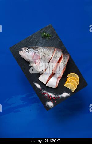 Nahaufnahme von frischem rohem Lachs, Forellenfilet mit Meersalz und Zitrone auf schwarzem Stein-Schneidebrett auf blauem Hintergrund Stockfoto