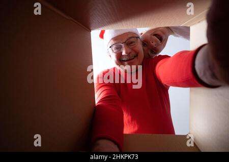 Senior Mann in weihnachtsmütze Auspacken, öffnen Karton und Blick nach innen. Git wird ausgepackt. Stockfoto