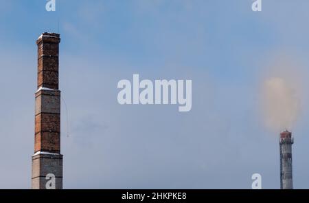 Alter Ziegelkamin auf verschwommenem Hintergrund von rauchigem Himmel und moderner Dampfleitung Stockfoto