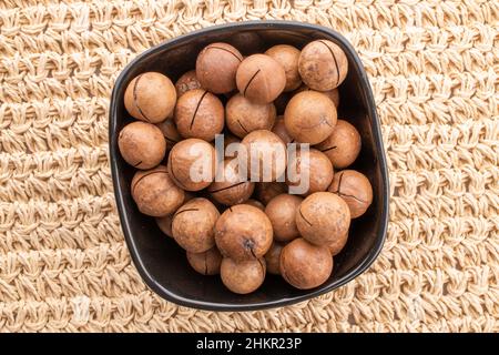 Mehrere reife braune Macadamianüsse in einer schwarzen Keramikschale, Nahaufnahme, auf einer Weinmatte, Draufsicht. Stockfoto