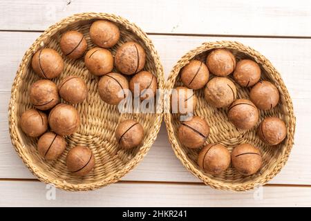 Mehrere reife braune Macadamianüsse in einer Strohschale, Nahaufnahme, auf einem Holztisch, Draufsicht. Stockfoto