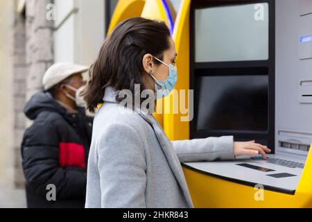 Menschen mit Gesichtsmaske im Freien, die an einem Geldautomaten Bankgeschäfte tätigen. Stockfoto