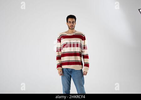 Auf weißem Grund steht der junge afroamerikaner mit Brille und gestreiftem Pullover. Stockfoto