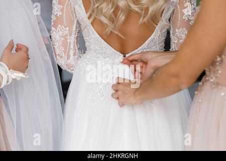 Wunderschöne, blonde Braut in einem weißen luxuriösen Kleid bereitet sich auf die Hochzeit vor. Vorbereitung am Morgen. Die Frau zieht sich ein Kleid an. Stockfoto