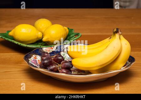 Früchte; Zitronen auf grünen Tellern, Trauben und Bananen auf einem antiken Teller auf einem Holztisch in der Küche Stockfoto