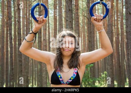 Porträt eines hübschen Teenagers, das vor dem Hintergrund eines Waldes auf Turnringen in der Natur aufzieht, Outdoor-Kurse Stockfoto