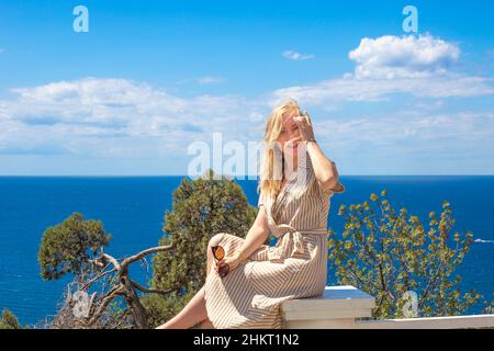 Junge schöne blonde Frau sitzt auf einem Hintergrund aus blauem Meer und blauem Himmel. Reisen und Tourismus. Stockfoto