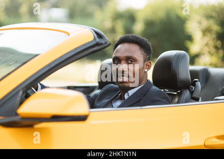 Seitenfenster des Fahrzeugs. Junge gutaussehende bärtige afrikanische Geschäftsmann Fahrer glücklich lächelnd Fahrsport gelben Auto. Hübscher junger Mann, der sich über sein neues Fahrzeug aufgeregt hat. Positiver Gesichtsausdruck Stockfoto