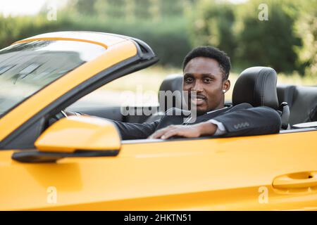 Seitenfenster des Fahrzeugs. Junge gutaussehende bärtige afrikanische Geschäftsmann Fahrer glücklich lächelnd Fahrsport gelben Auto. Hübscher junger Mann, der sich über sein neues Fahrzeug aufgeregt hat. Positiver Gesichtsausdruck Stockfoto