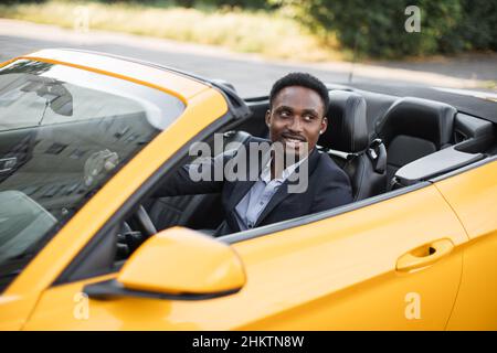 Seitenfenster des Fahrzeugs. Junge gutaussehende bärtige afrikanische Geschäftsmann Fahrer glücklich lächelnd Fahrsport gelben Auto. Hübscher junger Mann, der sich über sein neues Fahrzeug aufgeregt hat. Positiver Gesichtsausdruck Stockfoto