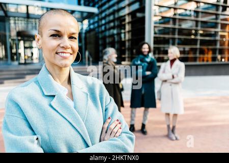 Glücklich lächelnde, professionelle Managerin mit gekreuzten Armen, die im Freien stehen Stockfoto