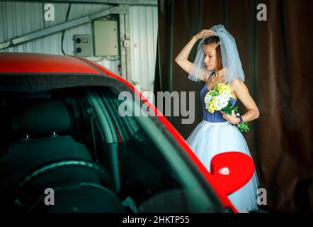Eine Frau in einem Schleier wäscht ein Auto bei einer Waschanlage. Hochzeitstag. Humor. Stockfoto