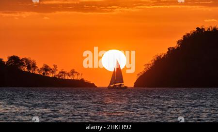 Vor der untergehenden Sonne und dem trüben orangenen Himmel zwischen zwei Landmassen vor der Küste Costa Ricas wird ein Segelboot mit vollen Segeln nach oben geschildet. Stockfoto